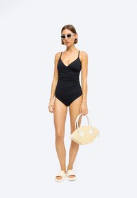 Costum de baie negru, cu un design pliat, bretele subțiri și decolteu în v. Modelul ține o geantă din nu puțin cu mână și poartă sandale albe cu talpă plată.