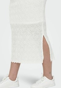 Witte kanten midi-rok met wervelpuntpatroon en zijsplit, gecombineerd met witte sneakers met een metallic accent. Golfdetail aan de onderrand.