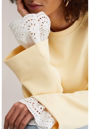 Mujer con una blusa de color amarillo pálido de mangas anchas y puños de encaje blanco, apoyando su rostro en su mano, mostrando un pendiente de aro dorado y cabello rizado.