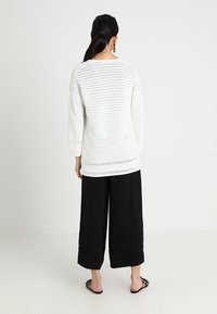 Vit stickad tröja med lös passform, försedd med öppna mönster och trekvartsärmar; tillsammans med svarta vida byxor.