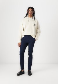 Krém színű pulóver kis logóval, sötétkék slim-fit nadrággal párosítva, fekete loaferekkel kiegészítve. A modell fehér háttér előtt áll.