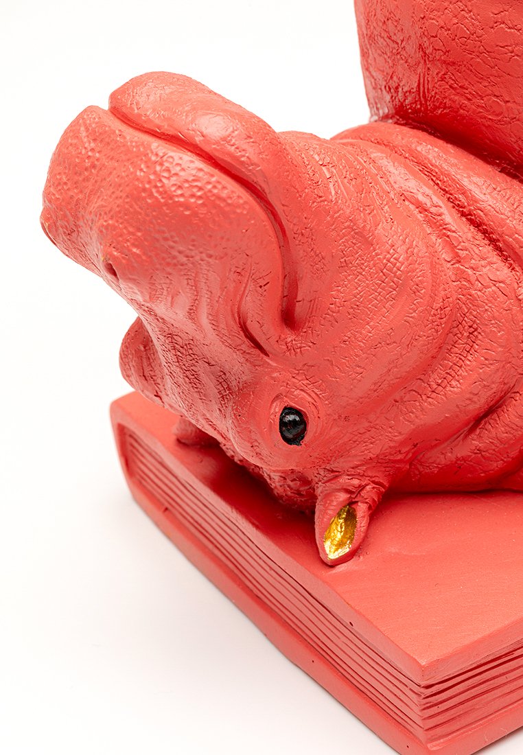 Rote Keramikschweinefigur, die auf dem Rücken liegt, auf einem geschlossenen Buch mit strukturierten Details und einem goldfarbenen Ohr.