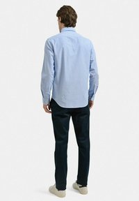 Ljust blå långärmad skjorta med knäppbar krage, rundad nederkant och en mjuk textur. Kombinerad med mörka byxor och beige skor.