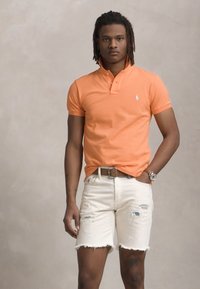 Oranžs polo krekls ar pogu apkakli, apvienots ar baltiem nolietotiem džinsa šortiem. Modelis valkā brūnu jostu un pulksteni.