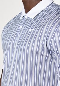 Camisa polo masculina de gola em cinza claro com riscas verticais azuis. Apresenta um discreto logótipo da Nike no peito e um tecido texturizado.