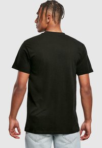 Black cotton T-shirt with short sleeves, a crew neck, and a relaxed fit. In the background, a person is viewed from the back, wearing light jeans.
