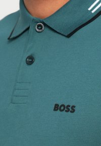 Türkizkék póló texturált felülettel, fekete gombokkal és fekete "BOSS" logóval. Gallérral rendelkezik, amely fekete és fehér díszítéssel van ellátva.