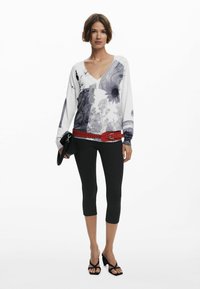 Sweter w serek w kolorze białym z graficznymi, czarno-szarymi wzorami kwiatowymi, w połączeniu z czarnymi legginsami w długości capri i czerwonym, ćwiekowanym paskiem.