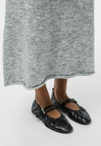 Bailarinas de cuero negro con dos correas y herrajes plateados, combinadas con un vestido de punto gris suave que presenta un dobladillo scalloped.