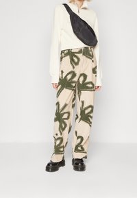 Personen bär en krämfärgad zip-up-tröja, beige byxor med stora gröna abstrakta blommönster, svarta kraftiga skor och en svart crossbody-väska.