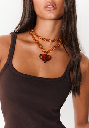  ravfarvet perlekæde med hjerteformet vedhæng, lagdelt med runde og ovale perler, båret over en brun tanktop.