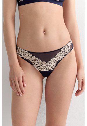 Ženský trup v námořnicky modrém spodním prádle ve stylu bikiny s béžovými květinovými krajkovými detaily, stojící na jednobarevném světle pozadí.