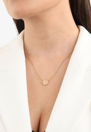 Collana d'oro con un pendente rotondo che presenta una superficie liscia e piccoli accenti decorativi; indossata sopra un blazer bianco e un top nero.