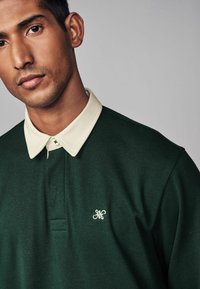 Zelena polo majica s svijetlosmešavim ovratnikom, koja sadrži mali izvezeni logo na prsima. Napravljena je od mekog materijala s glatkom teksturom.