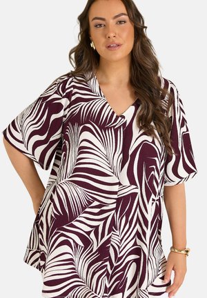 Kobieta z długimi brązowymi włosami, ubrana w luźną bluzkę z dekoltem w serek z bordowo-białym abstrakcyjnym wzorem liści oraz złotymi kolczykami kołami i bransoletką.