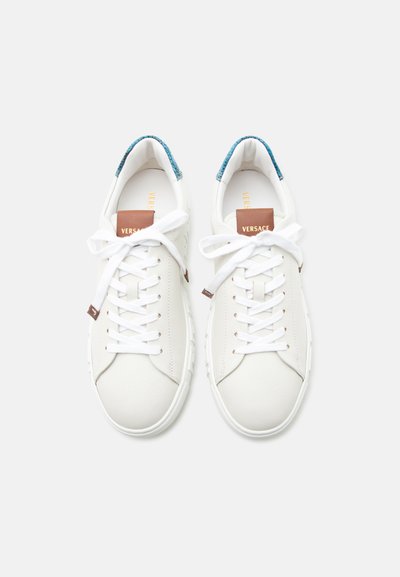 Baskets basses blanches avec des lacets blancs, des étiquettes de languette marron et des languettes de talon texturées bleues, vues de dessus sur un fond blanc.