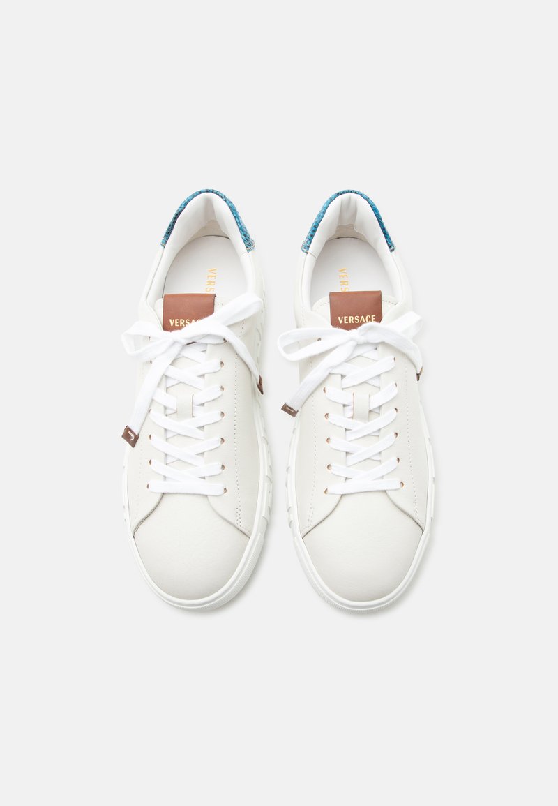 Baskets basses blanches avec des lacets blancs, des étiquettes de languette marron et des languettes de talon texturées bleues, vues de dessus sur un fond blanc.