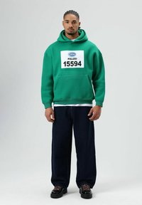 Groene hoodie met een voorzak en een wit label met de tekst "POLIZEI 15594." Gecombineerd met losse donkerblauwe spijkerbroek en zwarte schoenen met roze veters.