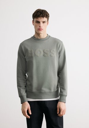 Mladý muž v sivom pulóvri s vystúpeným nápisom „BOSS“ a čiernych nohaviciach, stojaci pred jednoduchým bielym pozadím.