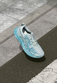 Chaussure de sport bleu clair avec une tige en maille texturée, un design motif et une semelle rembourrée, positionnée sur un pavé près d'un passage piéton.