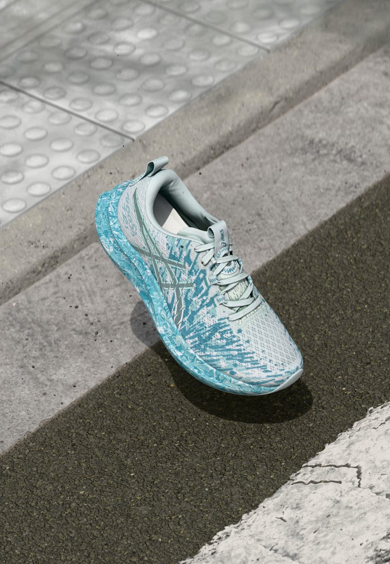 Chaussure de sport bleu clair avec une tige en maille texturée, un design motif et une semelle rembourrée, positionnée sur un pavé près d'un passage piéton.
