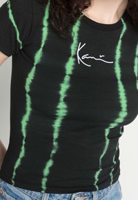 Black t-shirt featuring green tie-dye vertical stripes, with white embroidery of the word 'Kari' near the neckline. Soft cotton material.