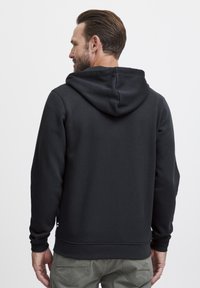 Haut à capuche noir avec une texture douce, comportant une capuche à cordon de serrage, des poignets côtelés et un ourlet droit. La vue de dos montre un tissu lisse et des lignes épurées.