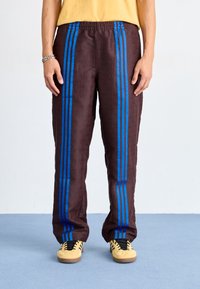 Pantaloni della tuta marroni con strisce blu, caratterizzati da un motivo testurizzato e una vita elastica. Abbinati a scarpe gialle, su uno sfondo blu.