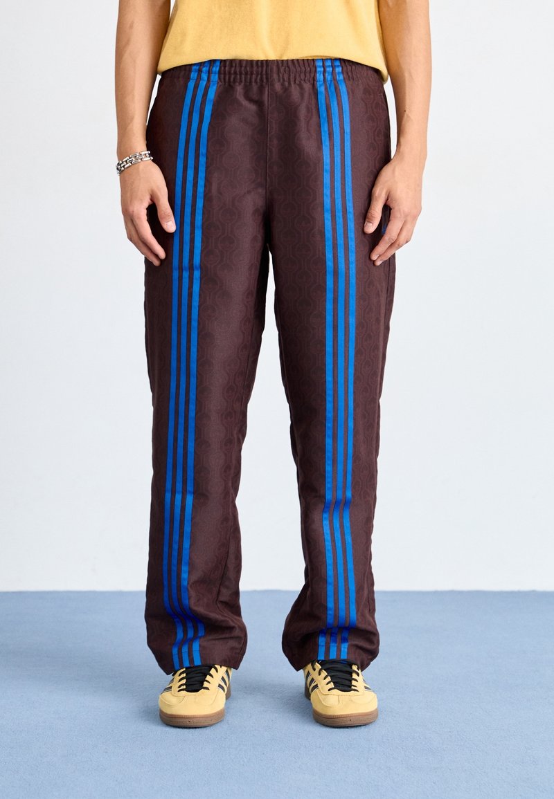 Pantaloni della tuta marroni con strisce blu, caratterizzati da un motivo testurizzato e una vita elastica. Abbinati a scarpe gialle, su uno sfondo blu.