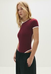 Bordo bodysuit s bijelim obrubom, kratkim rukavima i otvorenim stranama, u kombinaciji s crnim širokim hlačama. Glatka tekstura i oblikovani dizajn.