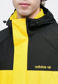 Μαύρο και κίτρινο μπουφάν Adidas με φερμουάρ στο μπροστινό μέρος, με μια μεγάλη κίτρινη ρίγα και λογότυπο στο στήθος. Λεία υφή υφάσματος.