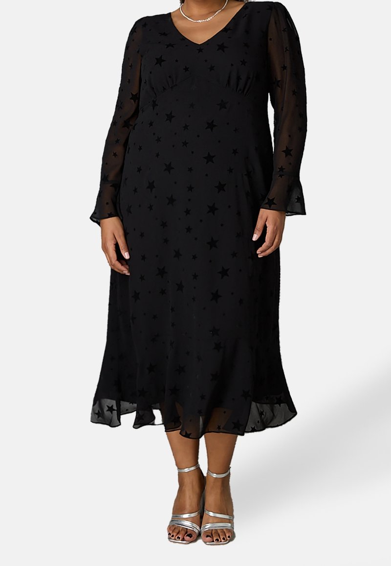 Musta mekko, jossa on läpinäkyvät pitkät hihat, tähtikuvio, V-kaula-aukko, A-linja ja levenevä helma. Yhdistetty hopeisiin remmisandaaleihin.