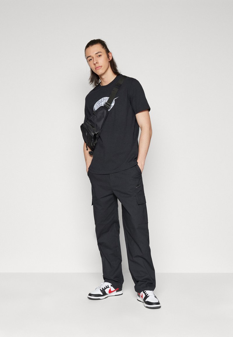 T-shirt noir avec un design graphique, pantalon cargo noir avec poches, et baskets blanches avec accents noirs et rouges.