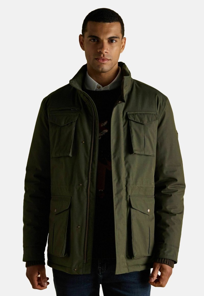 Ciemnozielona kurtka z wysokim kołnierzem, wieloma przednimi kieszeniami i zapięciem na zamek. Tkanina wydaje się teksturowana i lekko błyszcząca.