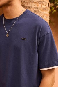 Marineblaues Baumwoll-T-Shirt mit kurzen Ärmeln; verfügt über ein kleines schwarzes Etikett mit Text. Geschichteter weißer Stoff an den Ärmelabschlüssen.