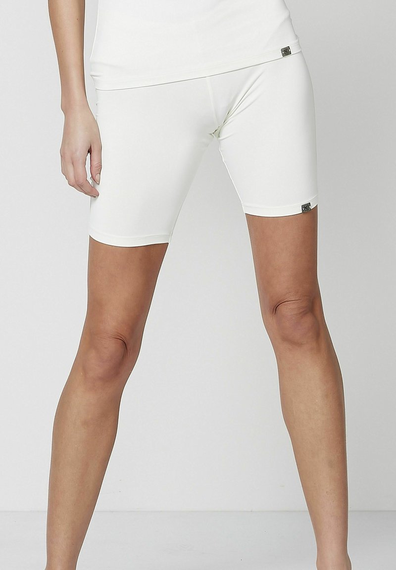 Femme portant un short blanc moulant mi-cuisse et un haut blanc assorti, debout, jambes légèrement écartées, sur un fond uni.