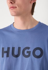 Mėlynas medvilninis marškinėliai su apvalia iškirpte, ant priekinės dalies didelėmis juodomis raidėmis parašyta "HUGO". Minkšta tekstūra ir kasdieniškas fasonas.