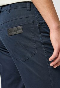Pantalones de mezclilla azul marino con un bolsillo trasero que presenta un parche de cuero negro con el logo de Wrangler y un patrón de costura distintivo. Textura suave.