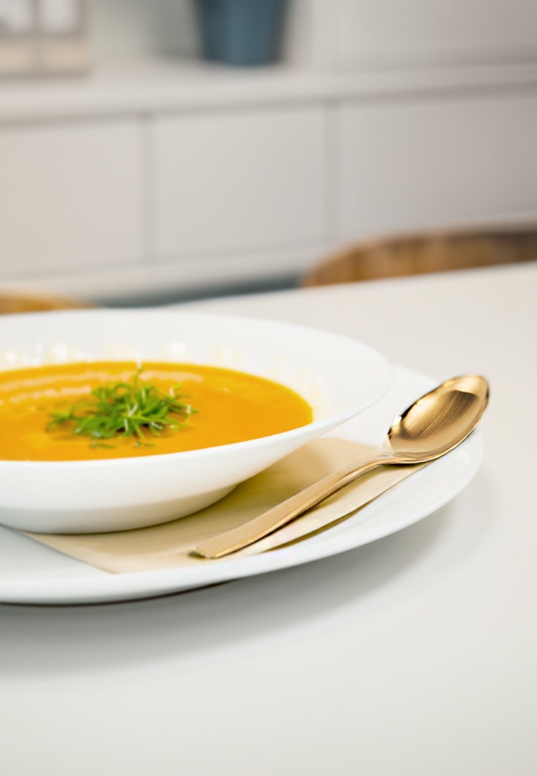 Weißer Schüssel gefüllt mit orangefarbener Suppe, garniert mit grünen Kräutern, begleitet von einem goldenen Löffel auf einer cremefarbenen Serviette auf einem weißen Teller.