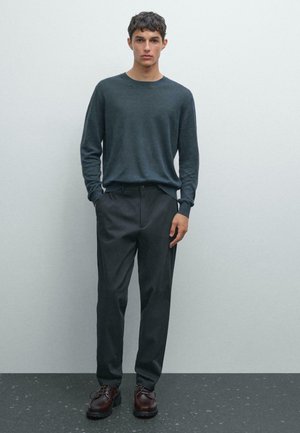 Young man standing, wearing a dark gray long-sleeve sweater, dark trousers, and brown leather lace-up shoes against a plain gray background.