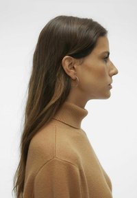 Bež turtleneck pulover iz mehkega materiala, ki ima rebrast ovratnik in šive. Model z dolgimi rjavimi lasmi in uhani v obliki obroča gleda levo.