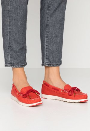 Jambes portant un jean gris retroussé et des chaussures style mocassins rouges avec des semelles blanches épaisses et des détails de nœud sur le dessus.