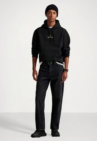 Camisola com capuz preta com logótipo dourado, usada sobre camisa branca. Jeans escuros, sapatilhas pretas e gorro preto completam o conjunto. Posição em pé.