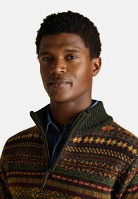 Kötött pulóver cipzáros gallérral, zöld alapszínnel és sárga, piros és barna színű, sokszínű geometrikus mintákkal. Texturált gyapjú anyag.