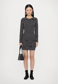 Black long-sleeve dress with white polka dots, fitted silhouette, and gathered neckline. Accessories include a small black handbag and shoes with accents.