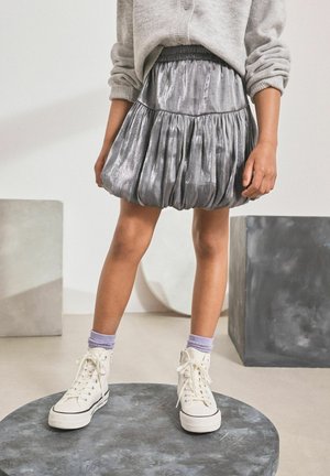 Jupe évasée en métal gris avec des plis, taille élastique. Associée à des baskets montantes blanches et des chaussettes lavande, debout sur un socle rond en pierre.