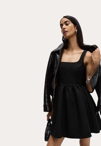 Abito nero con scollatura quadrata, gonna svasata e tessuto strutturato, abbinato a una giacca di pelle nera lucida e una piccola borsa.