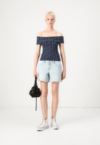 Mørkeblå avskåren topp med hvitt blomsterprint, lyseblå denimshorts og hvite høyhælte joggesko; holder en svart blomstertrukket veske.