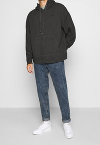 Felpa nera oversize con tasca frontale, abbinata a jeans blu chiaro e sneakers bianche. La felpa presenta un cappuccio con lacci e dettagli cuciti.