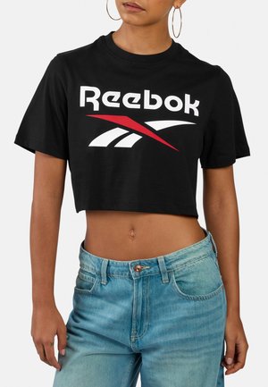 Person trägt ein schwarzes, bauchfreies Reebok-T-Shirt mit rotem und weißem Logo, hellblaue hoch taillierte Jeans und große Creolen-Ohrringe.